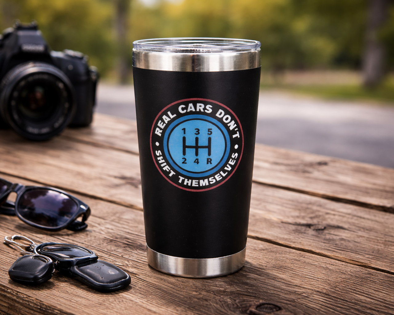 Black tumbler with a logo on a wooden table with sunglasses and keys, blurred camera in the background