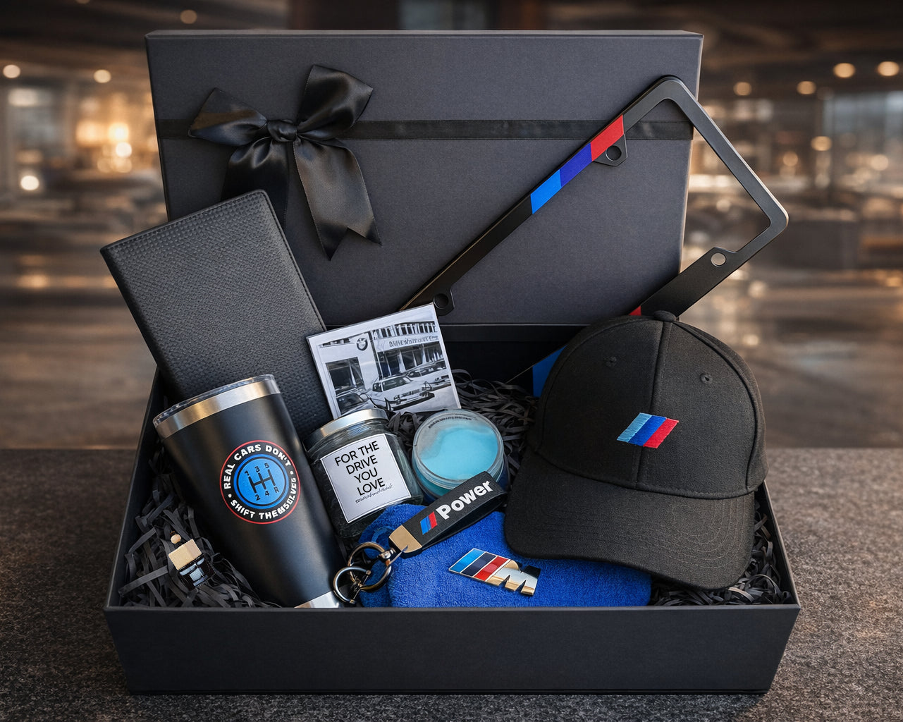 Gift box with various items including a tumbler, cap, and keychain on a blurred background.