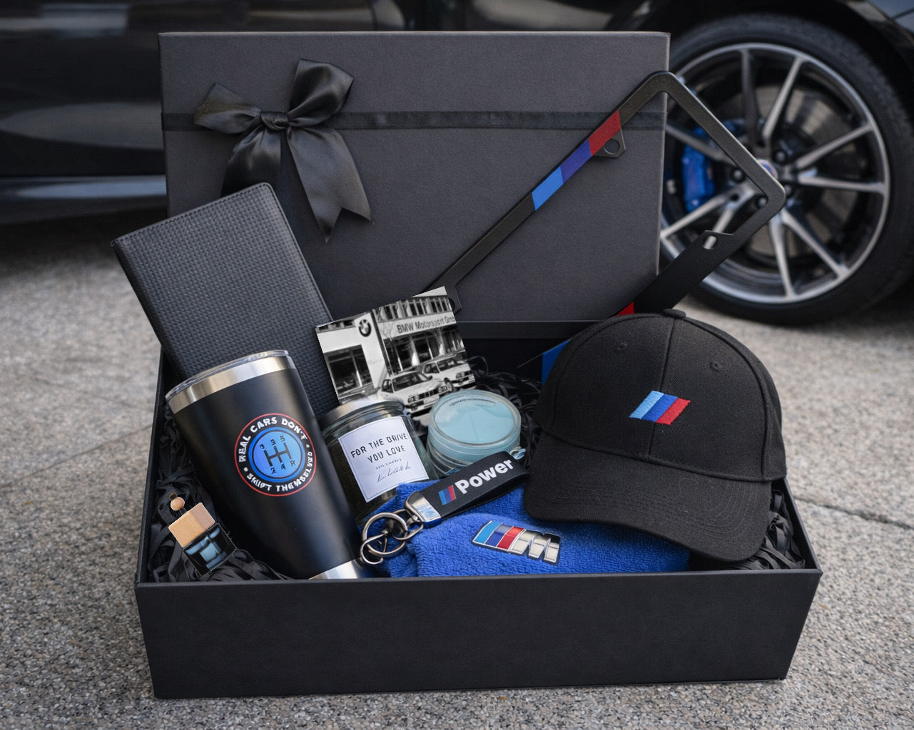 Gift box with car-themed items including a tumbler, cap, and keychain on a concrete surface.