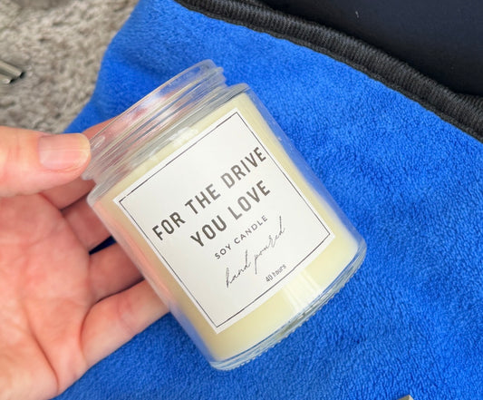 Hand holding a small candle labeled 'For the Drive You Love' on a blue surface.